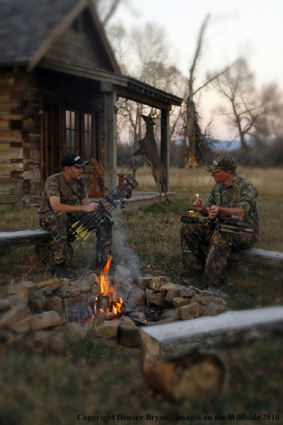 Archery hunters sitting around campfire with old hunting shack in background. (Original image # 11047-001.34D)