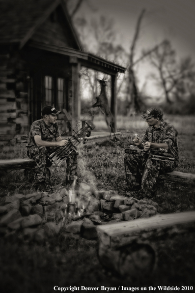Archery hunters sitting around campfire with old hunting shack in background. (Original image # 11047-001.34D)