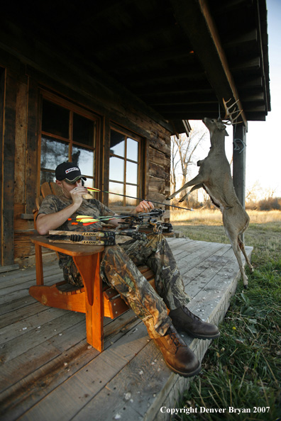 Archery hunter sittting on porch of old hunting shack where bagged white-tail hangs