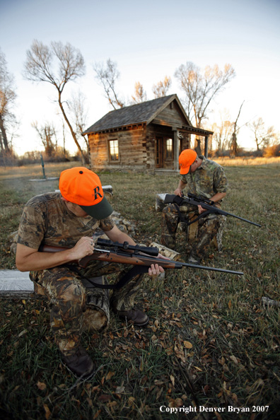 Hunters sitting around campfire in front of an old hunting shack where a white-tailed deer hangs.