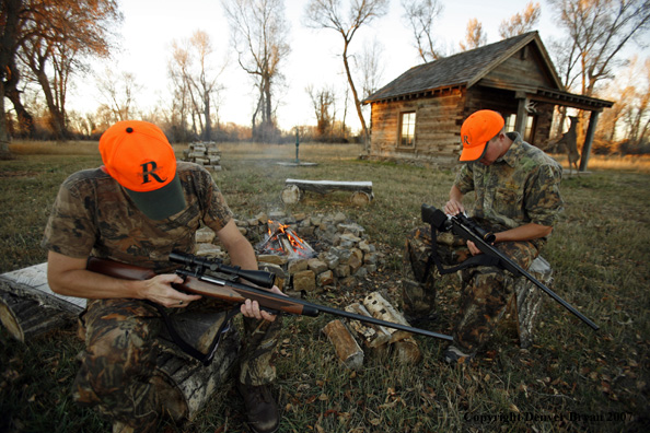 Hunters sitting around campfire in front of an old hunting shack where a white-tailed deer hangs.