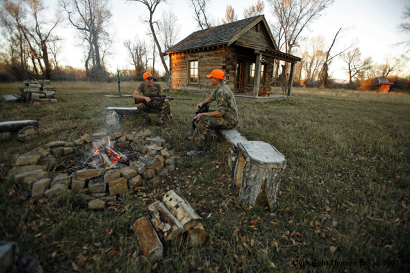 Hunters sitting around campfire in front of an old hunting shack where a white-tailed deer hangs.