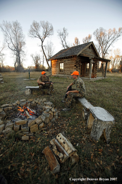 Hunters sitting around campfire in front of an old hunting shack where a white-tailed deer hangs.