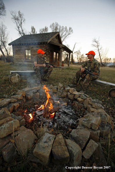 Hunters sitting around campfire in front of an old hunting shack where a white-tailed deer hangs.