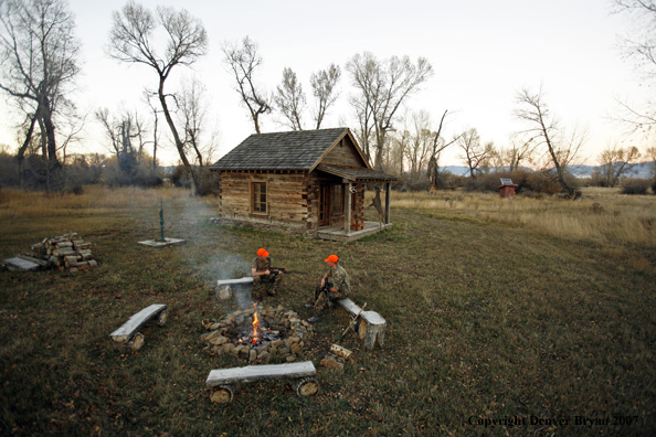 Hunters sitting around campfire in front of an old hunting shack where a white-tailed deer hangs.