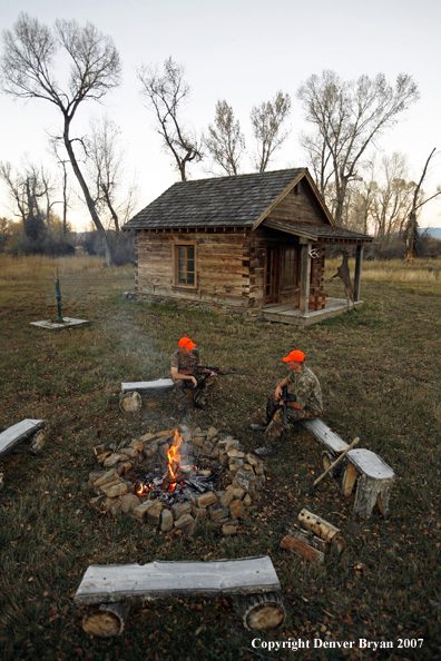 Hunters sitting around campfire in front of an old hunting shack where a white-tailed deer hangs.