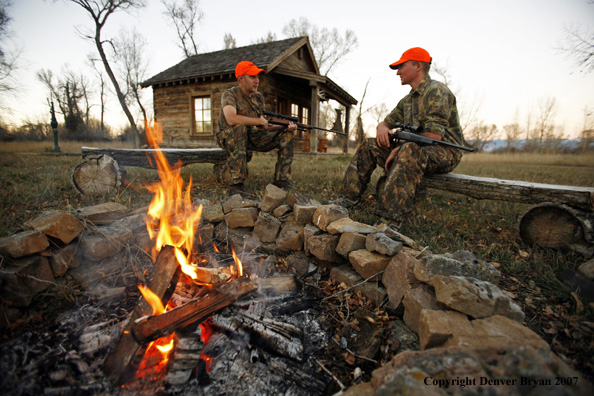 Hunters sitting around campfire in front of an old hunting shack where a white-tailed deer hangs.