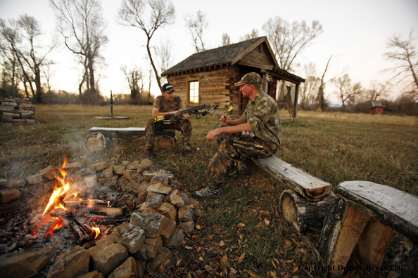 Archery hunters sitting around campfire with old hunting shack in background.