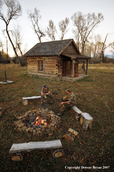 Archery hunters sitting around campfire with old hunting shack in background.