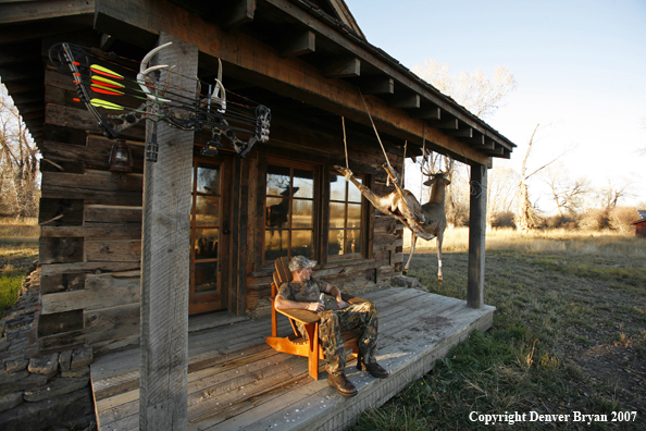 Archery hunter sittting on porch of old hunting shack where bagged white-tail hangs
