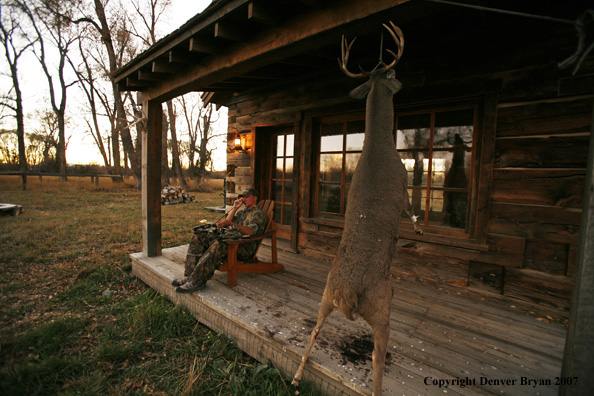 Archery hunter sittting on porch of old hunting shack where bagged white-tail hangs
