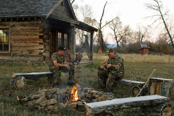 Archery hunters sitting around campfire with old hunting shack in background.