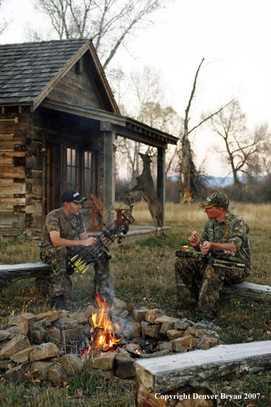 Archery hunters sitting around campfire with old hunting shack in background.