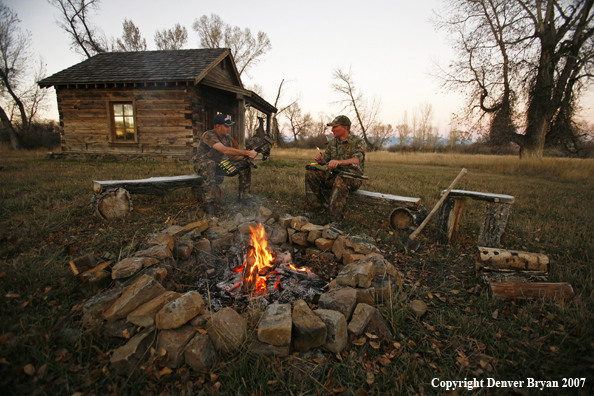 Archery hunters sitting around campfire with old hunting shack in background.