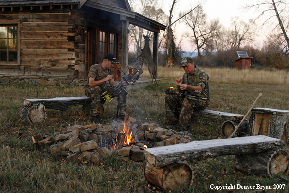 Archery hunters sitting around campfire with old hunting shack in background.