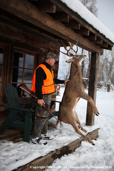 White-tailed deer hunter stands with buck hanging from cabin.