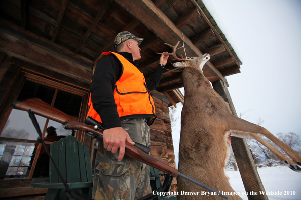 White-tailed deer hunter stands with buck hanging from cabin.