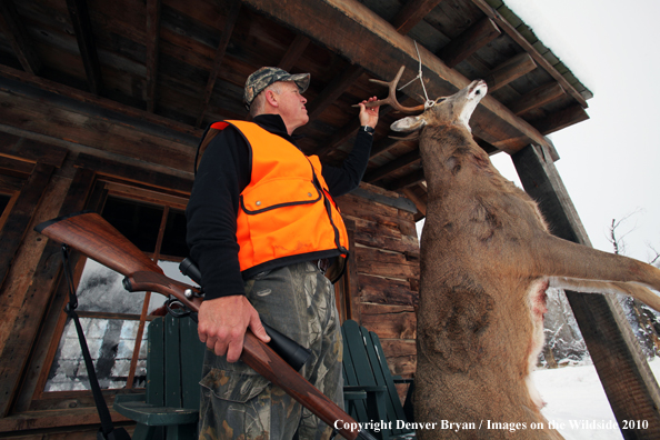 White-tailed deer hunter stands with buck hanging from cabin.