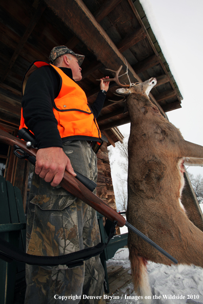 White-tailed deer hunter stands with buck hanging from cabin.
