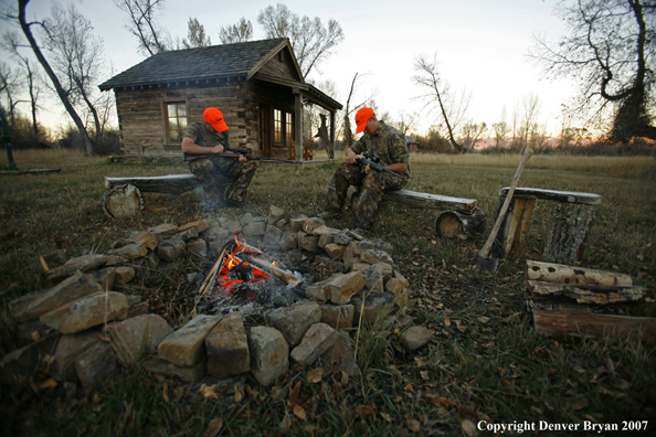 Hunters sitting around campfire in front of an old hunting shack where a white-tailed deer hangs.