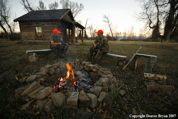 Hunters sitting around campfire in front of an old hunting shack where a white-tailed deer hangs.