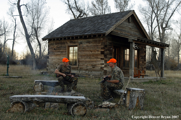 Hunters sitting around campfire in front of an old hunting shack where a white-tailed deer hangs.