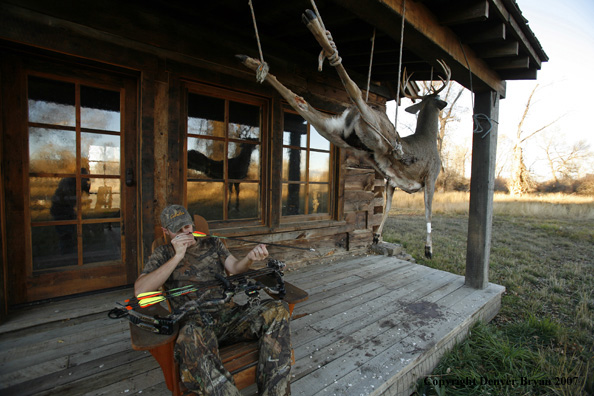 Archery hunter sittting on porch of old hunting shack where bagged white-tail hangs