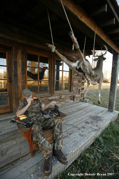 Archery hunter sittting on porch of old hunting shack where bagged white-tail hangs